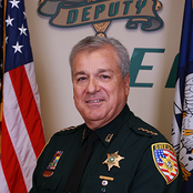 Man in sheriff's uniform, smiling, with flags and badge in the background.