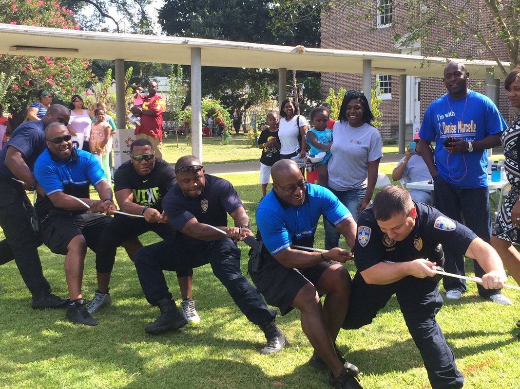 Men in blue shirts and black pants playing tug-of-war on grass, people watching in background.