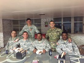 Group of soldiers in uniform pose at a table, some smiling and eating. Indoor setting, likely a break room.