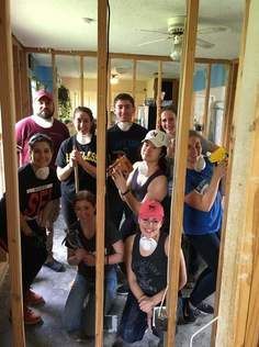 Group of people inside a framed room, some wearing masks, holding tools, smiling; construction scene.