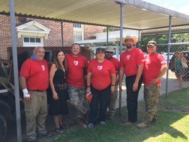 Group of people in red shirts pose outdoors under a canopy. Some wear hats, and one woman wears a black dress.