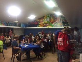 People seated at tables in a room, some wearing FEMA Corps jackets, eating and talking. Colorful murals on the wall.