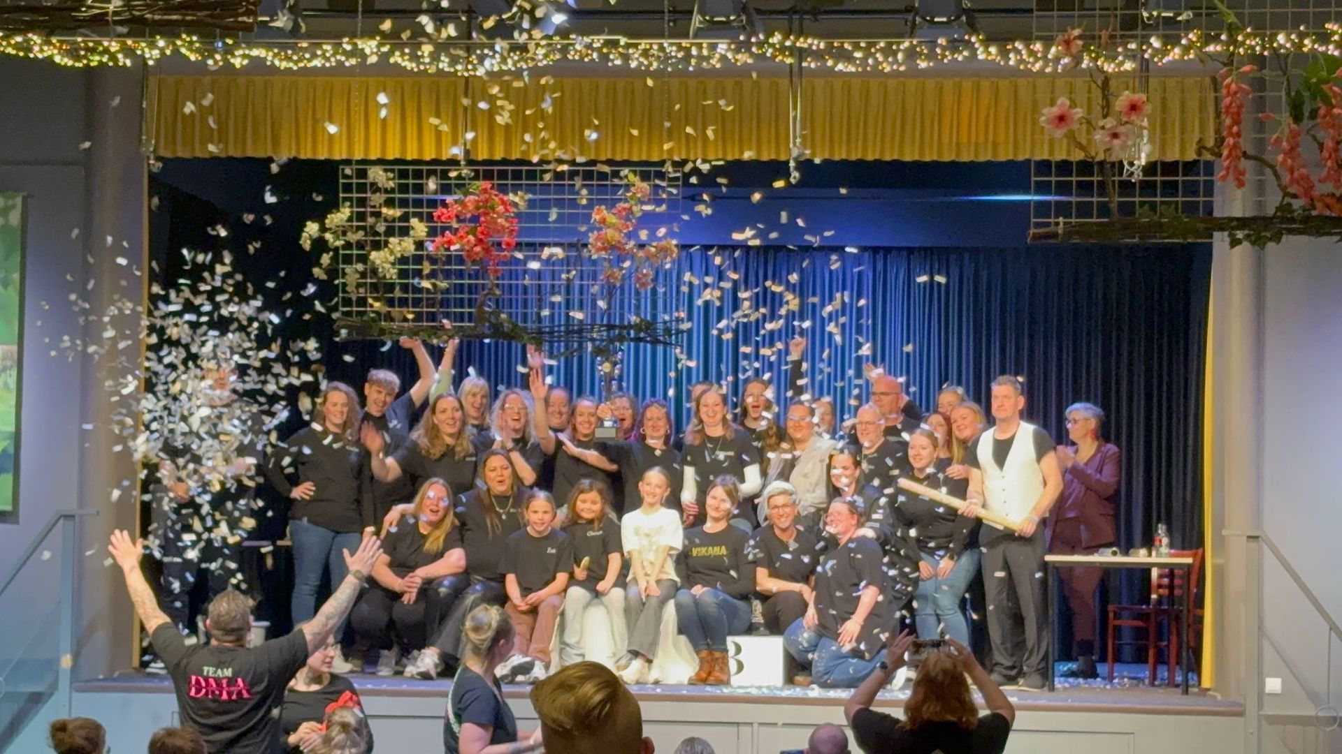 Een groep mensen viert feest op een podium te midden van neervallende confetti, waarbij sommigen juichen en lachen.