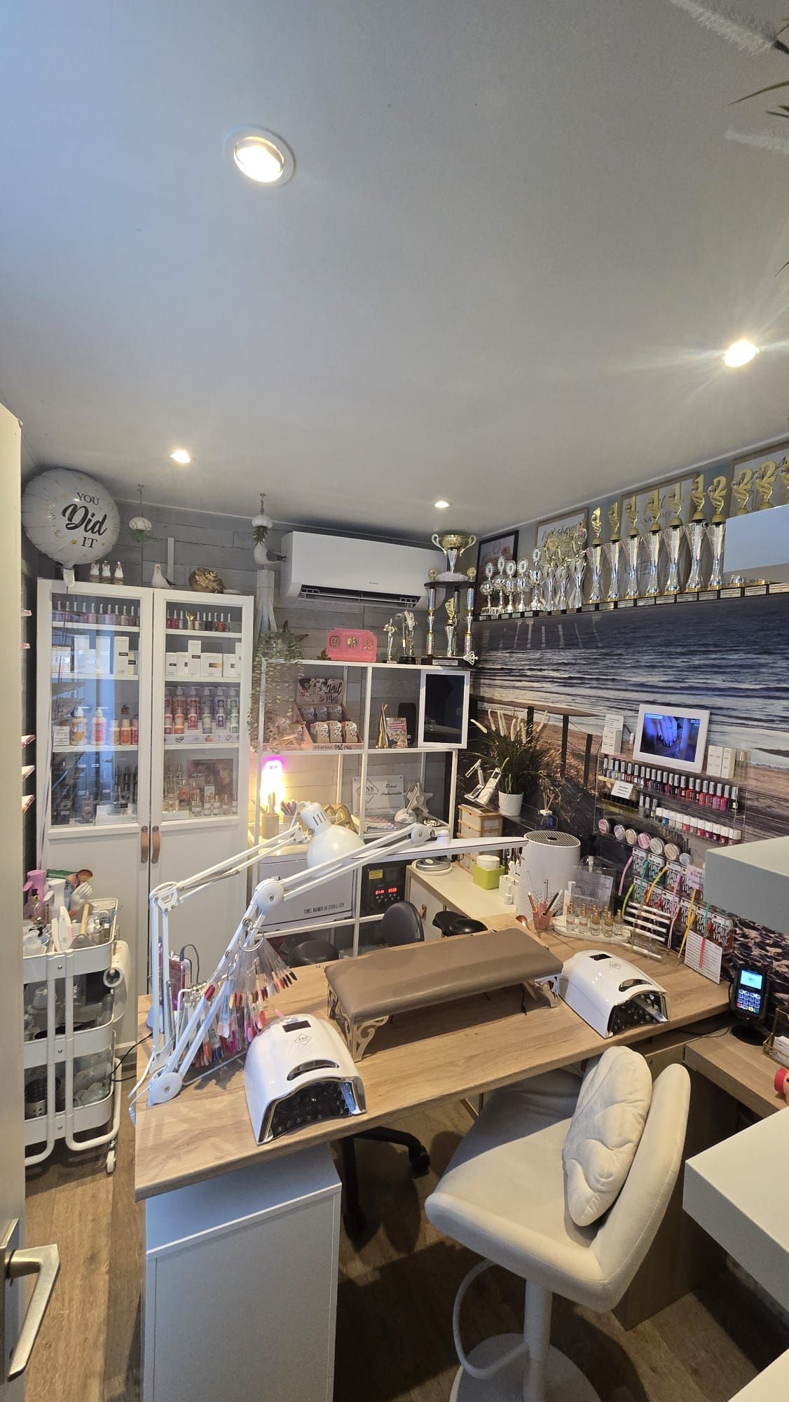 Een lichte werkplek voor nagelstylisten met een houten bureau, witte stoel, opbergplanken en bureaulampen in een studio-achtige omgeving.