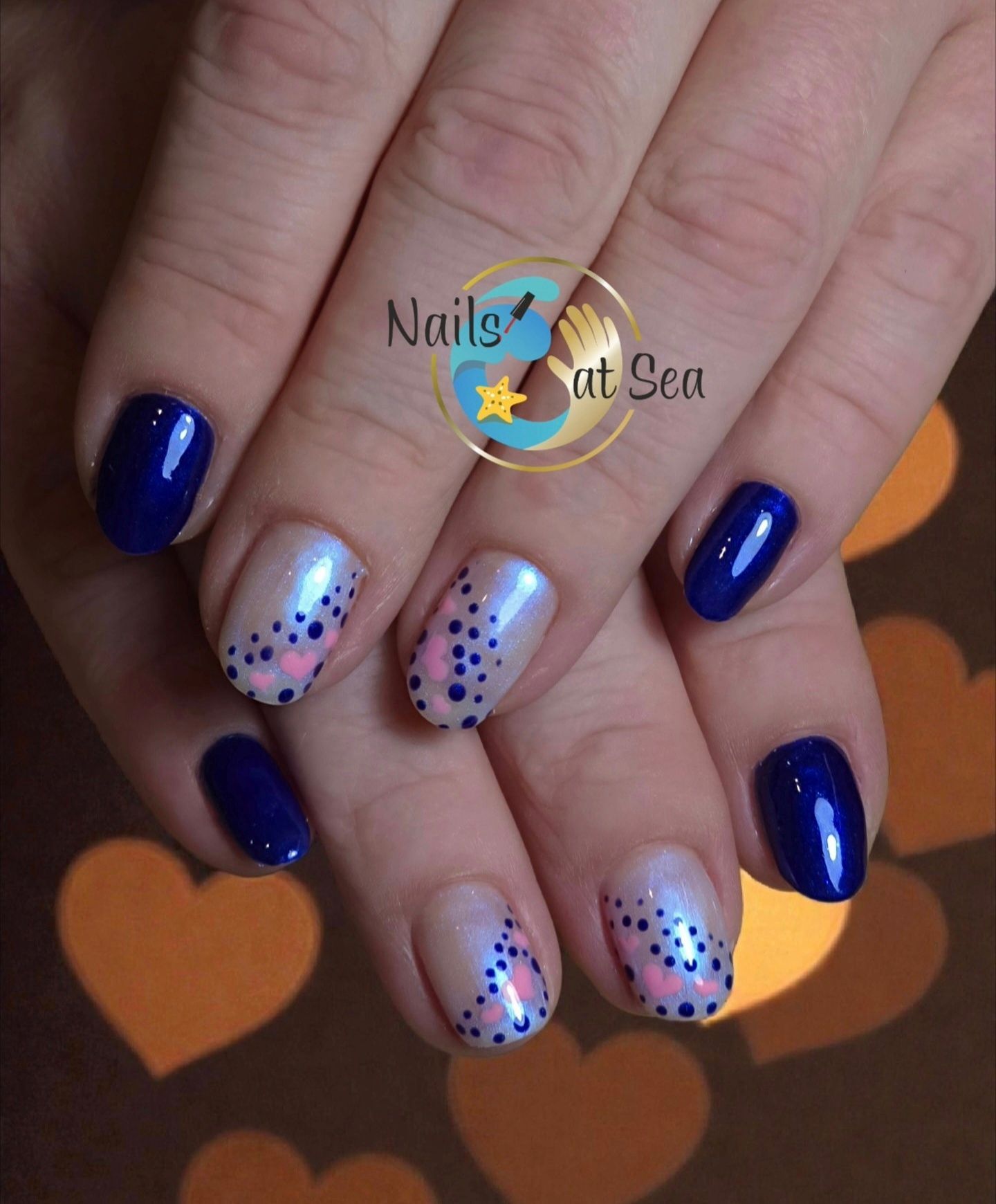 Verzorgde nagels met een glanzende, diepblauwe lak afgewisseld met een parelmoerachtige basis, versierd met blauwe en roze stippen.