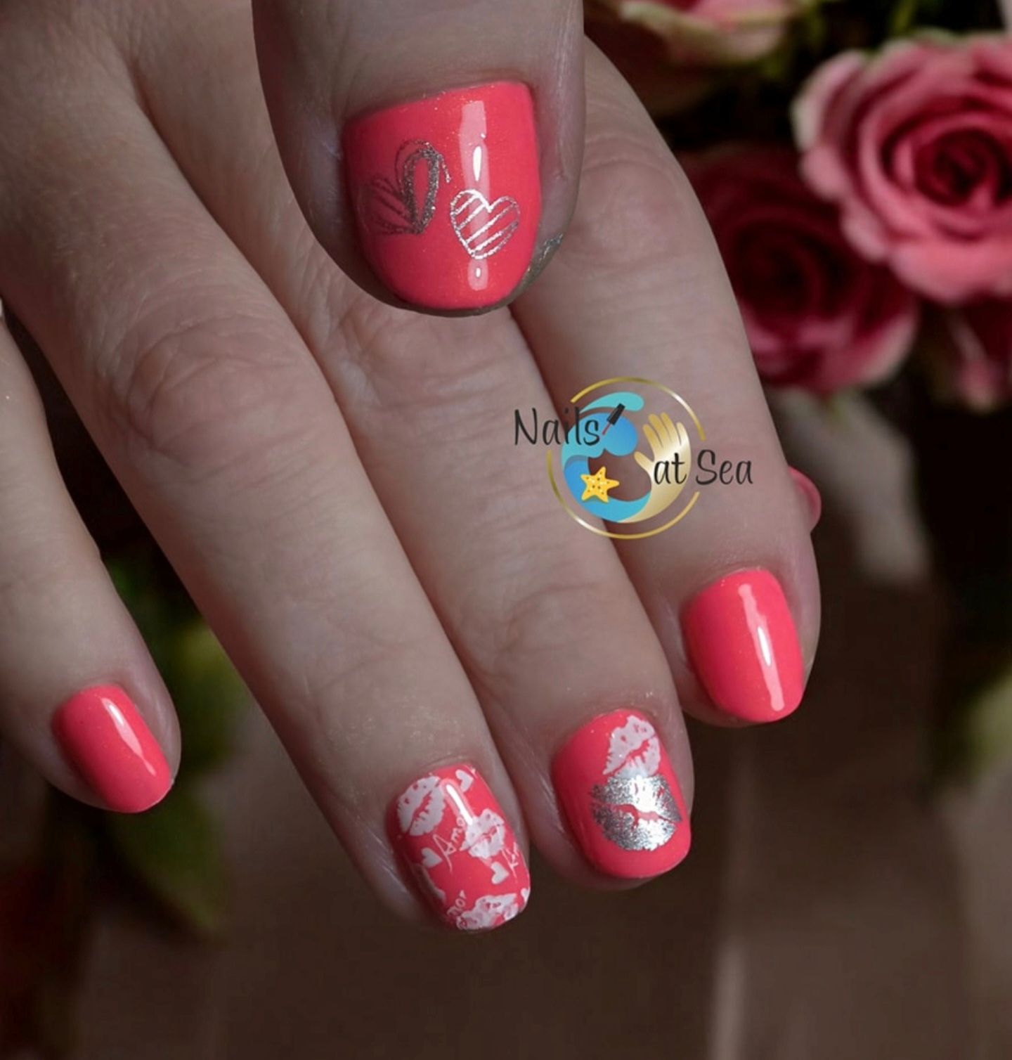 Close-up van fel koraalkleurige nagels met zilveren hartjes en lippenversieringen, naast wazige roze rozen.