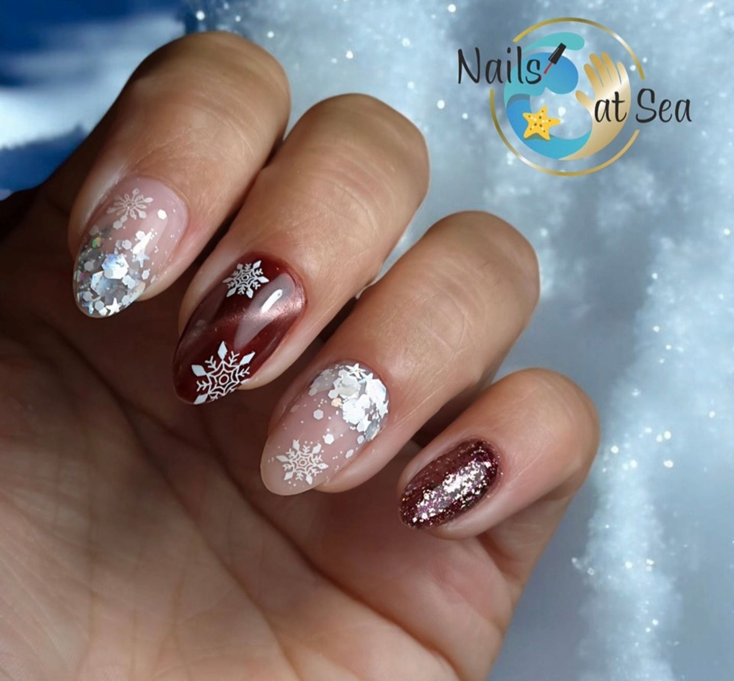 Een hand toont een manicure met winterthema, bestaande uit donkerrode cat-eye nagellak, zilveren glitter en witte sneeuwvlokjes.