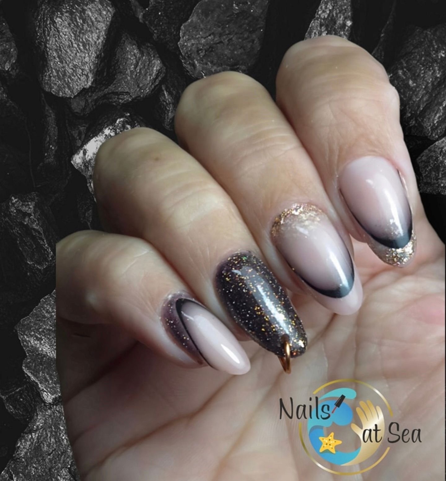Verzorgde amandelvormige nagels met een zwart-nude kleurverloop, gouden glitteraccenten en een klein metalen bedeltje.