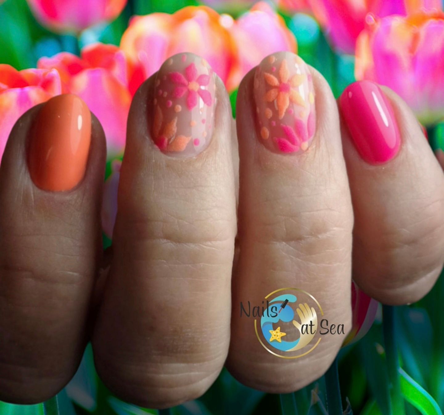Vier vingernagels met een lentemanicure in oranje, roze en bloemmotieven tegen een wazige tulpenachtergrond.