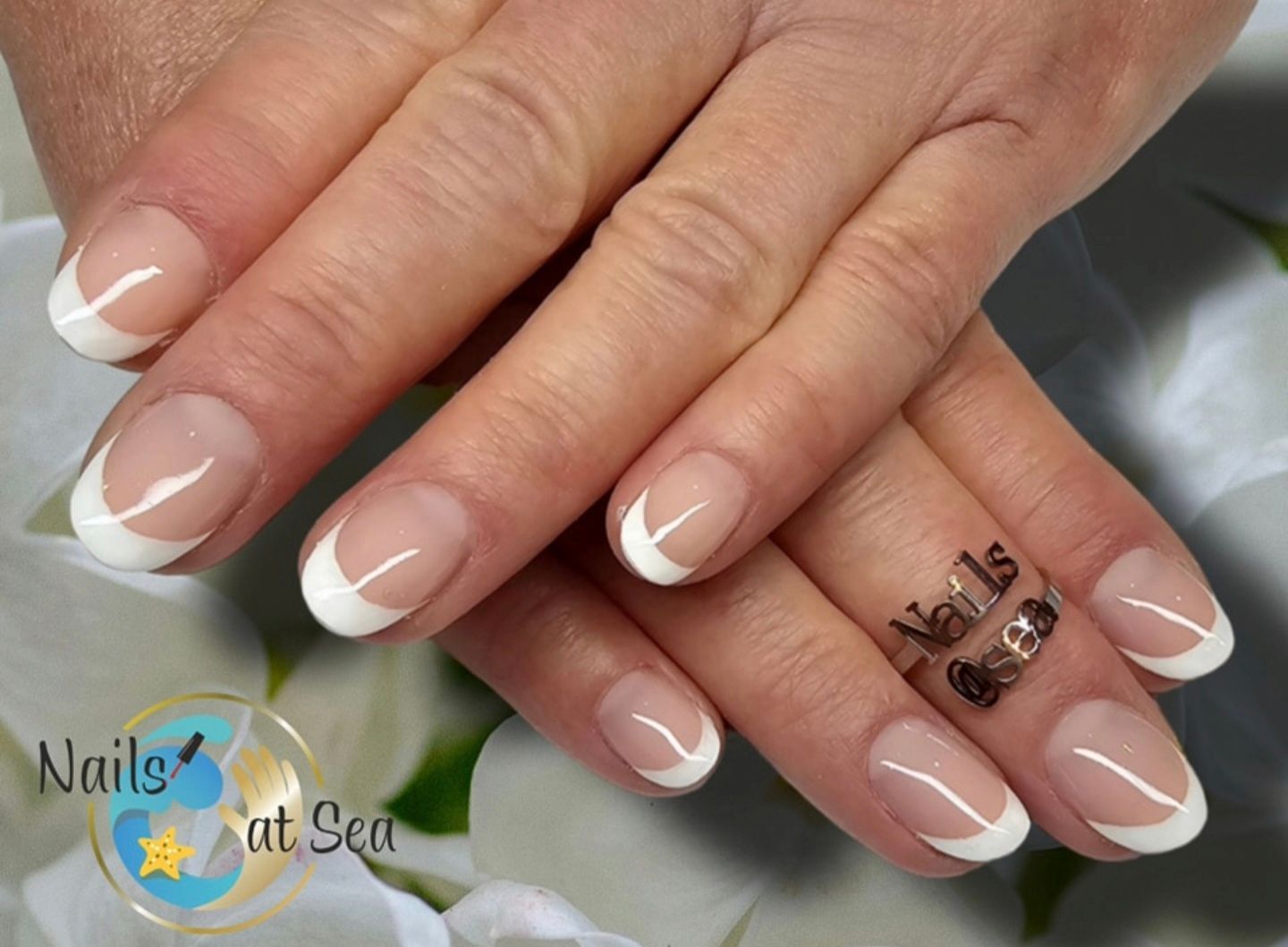 Een close-up van handen met een klassieke Franse manicure met strakke witte nagelpunten, tegen een zachte bloemenachtergrond.