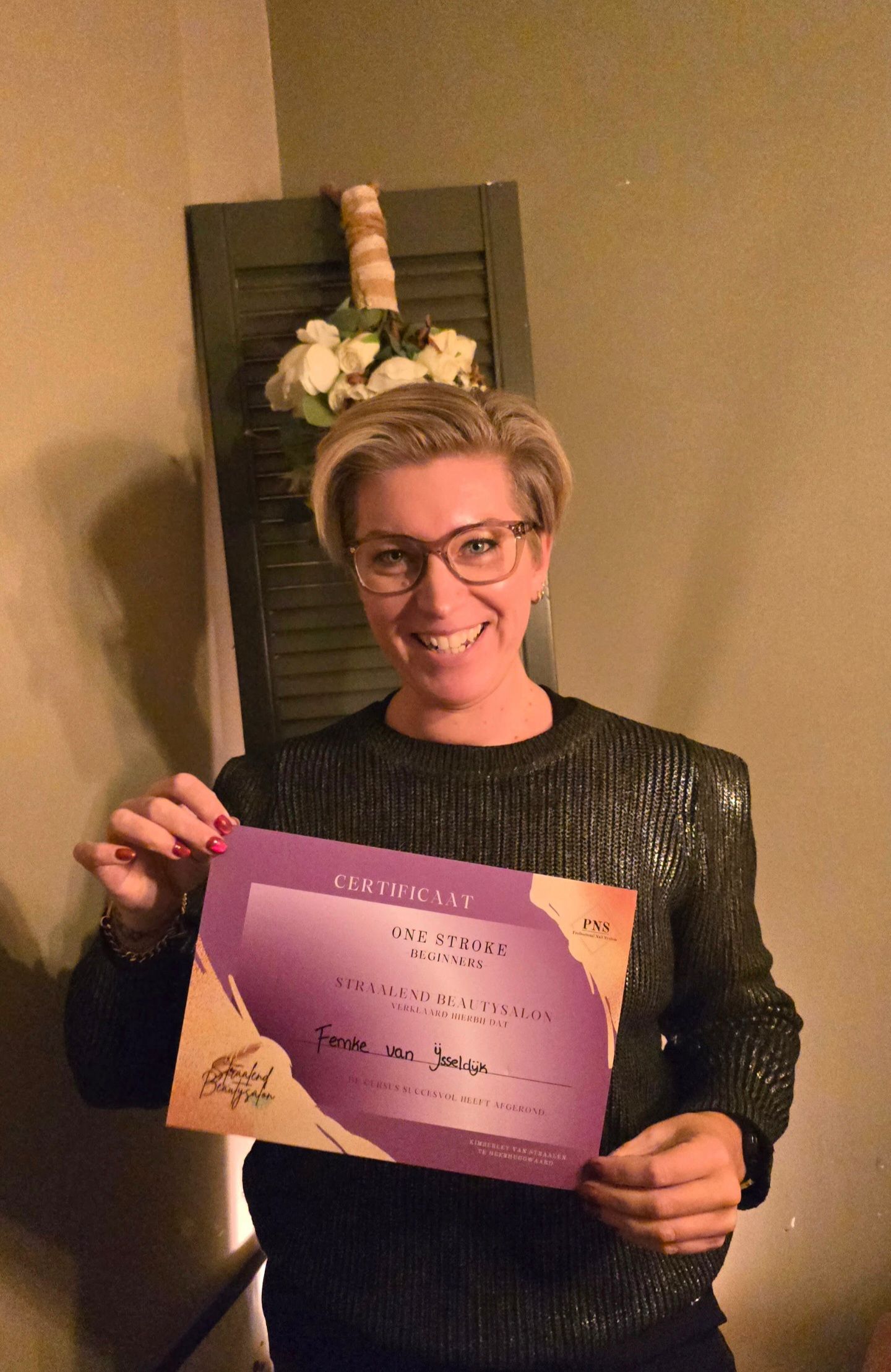 Een glimlachend persoon in een donkere trui houdt binnenshuis een paars rechthoekig certificaat omhoog.