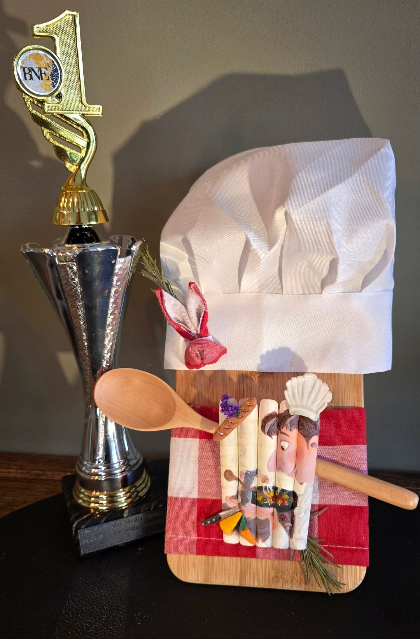 Naast een houten snijplank, versierd met een koksmuts, een rood geruit tafelkleed en een lepel, staat een trofee voor de eerste plaats.