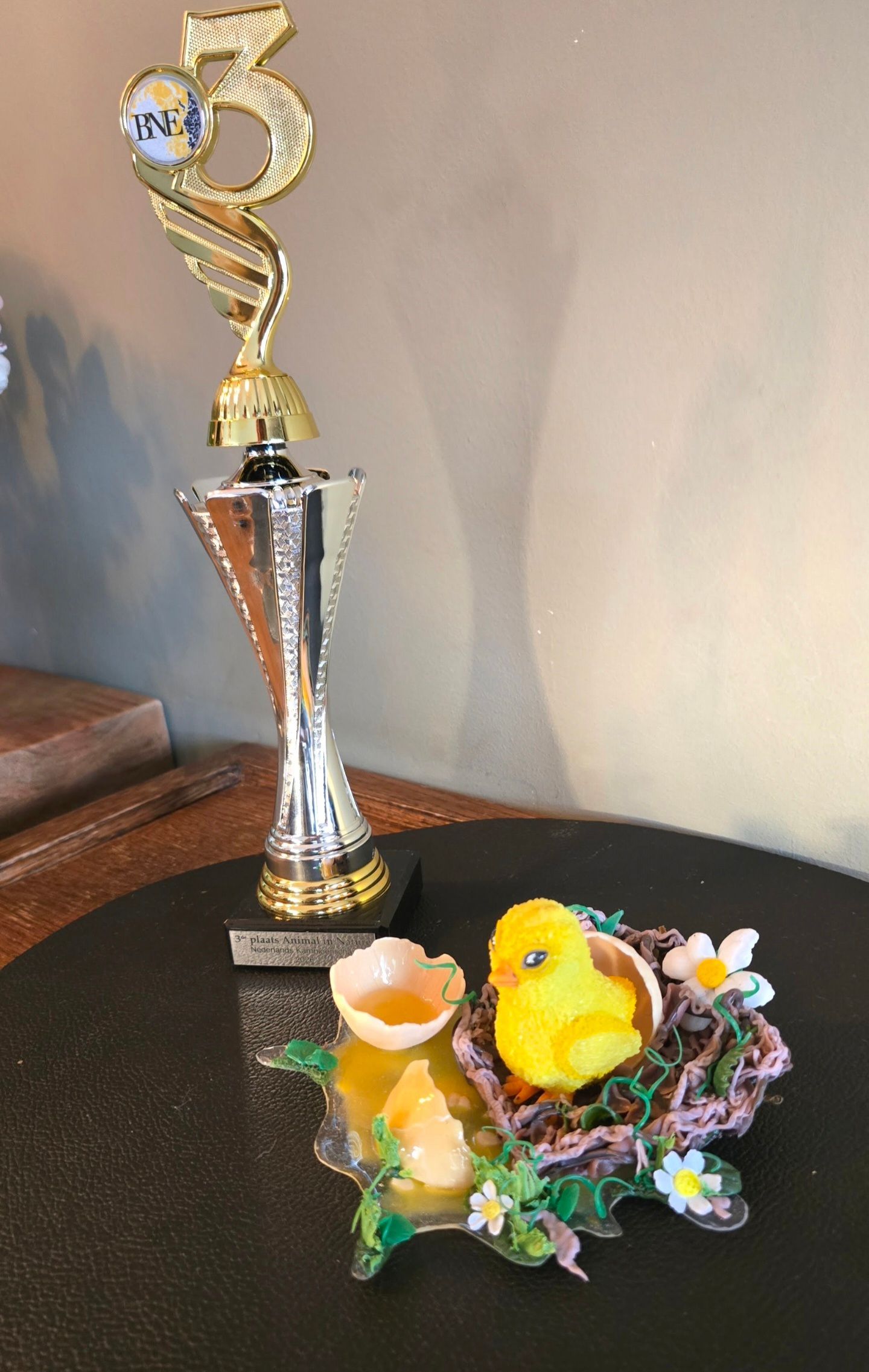 Naast een gouden en zilveren trofee staat een klein, geel speelgoedkuikentje in een decoratief nestje met bloemen op een donkere ondergrond.