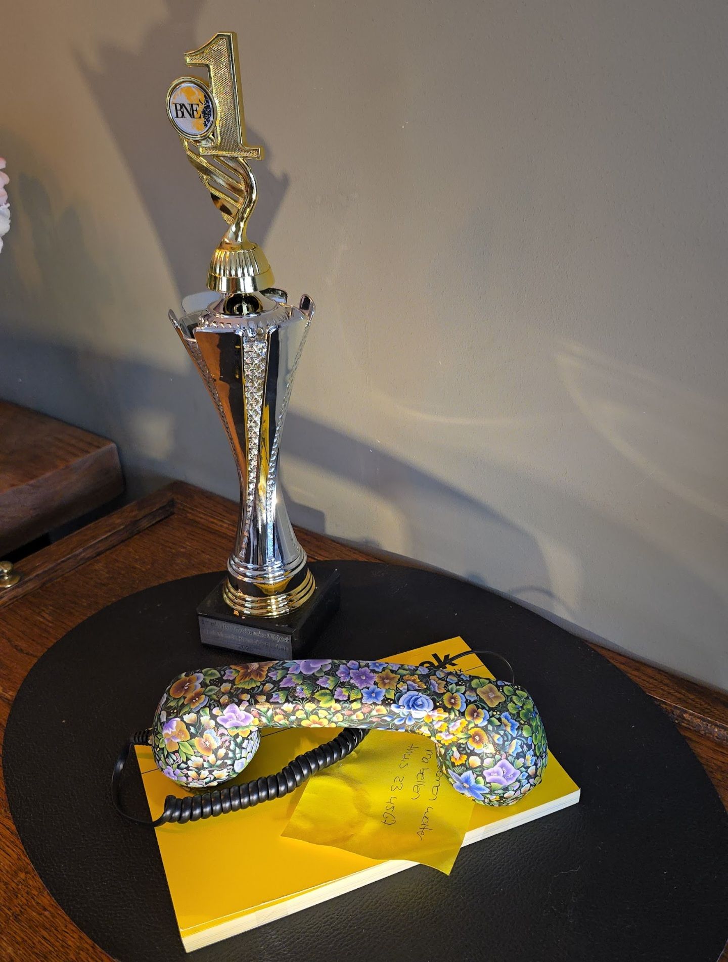 Een hoge trofee en een telefoonhoorn met bloemenmotief rusten op een geel notitieblok, alles geplaatst op een donkere, ronde ondergrond.