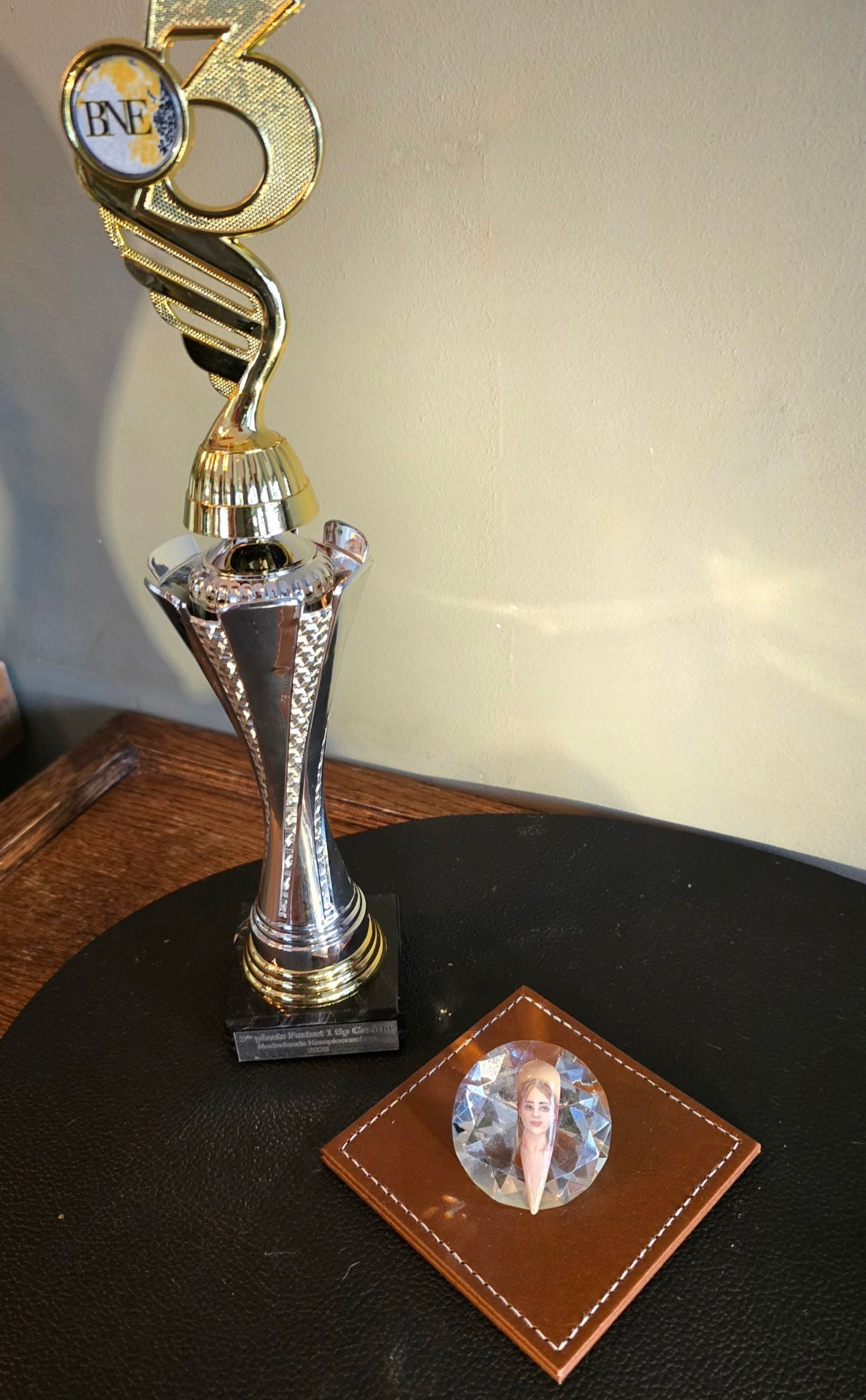 Een gouden en zilveren trofee staat op een donkere ondergrond naast een vierkante leren onderzetter met een klein rond kristallen ornament.