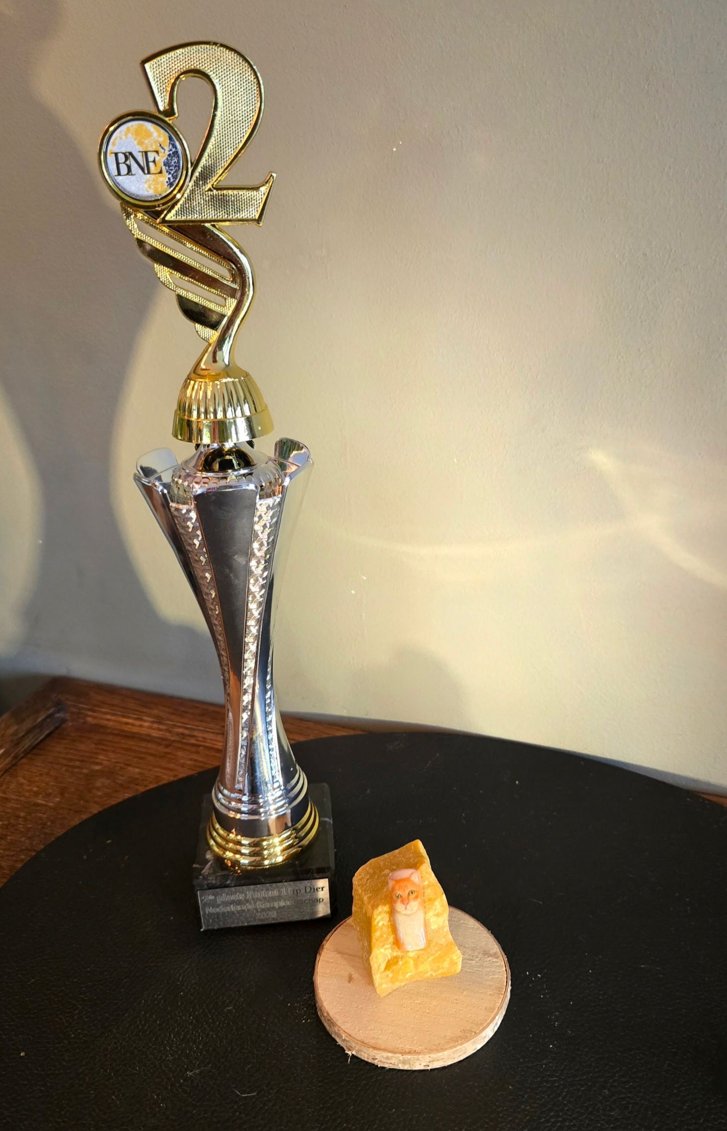 Een trofee voor de tweede plaats naast een klein, geel, vierkant snoepje met amandel erop, op een rond crackertje, op een zwarte ondergrond.