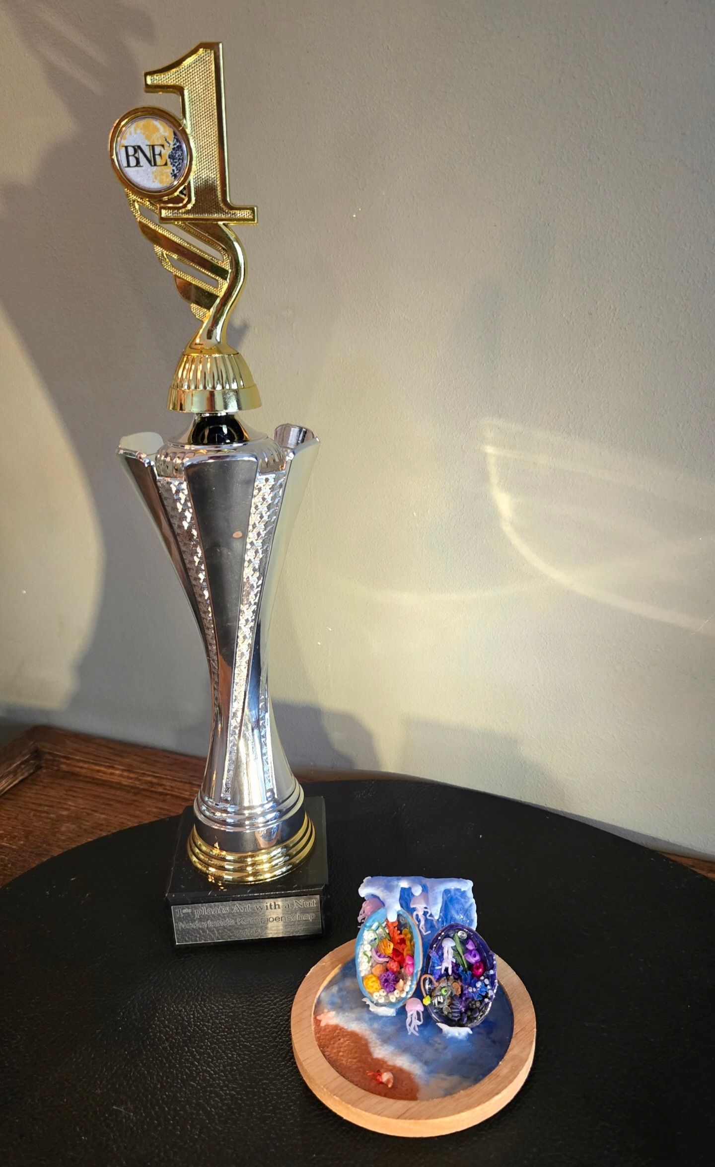 Naast een kleine, ronde houten onderzetter, versierd met kleurrijke strandkunst, staat een gouden en zilveren trofee voor de eerste plaats.