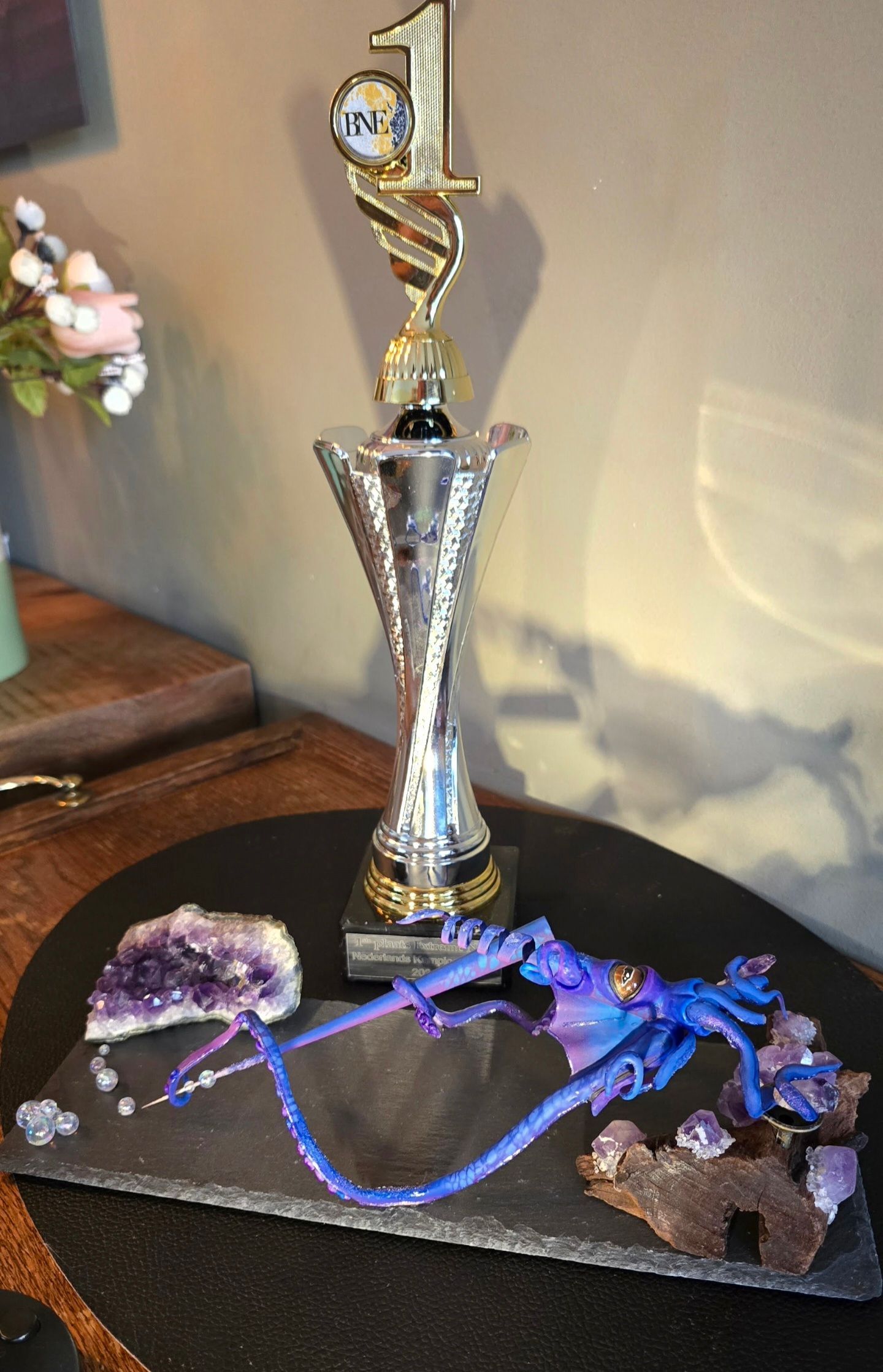 Een gouden en zilveren trofee voor de eerste plaats staat achter een paarse draak, die op een leistenen sokkel met amethistkristallen staat.