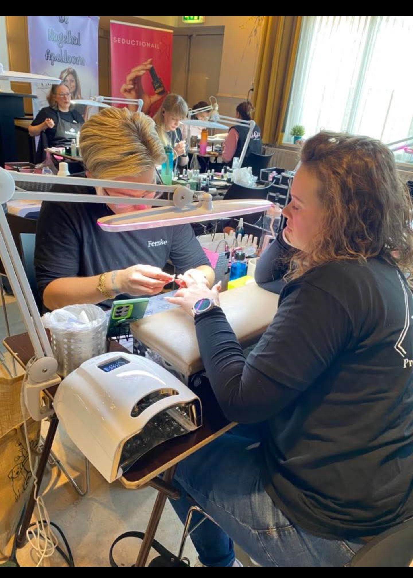 Een nagelstyliste voert een manicure uit op de hand van een klant aan een bureau, verlicht door een lamp in een helder verlichte studio.