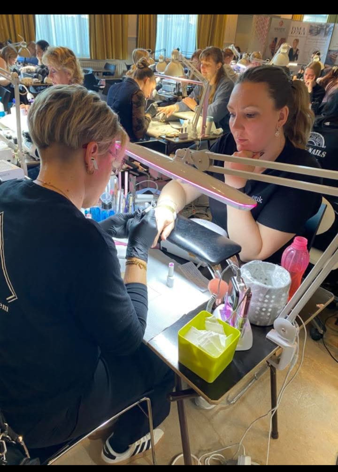 Een nagelstyliste met zwarte handschoenen voert een manicure uit op de hand van een klant in een drukke werkplaats met meerdere andere werkplekken.