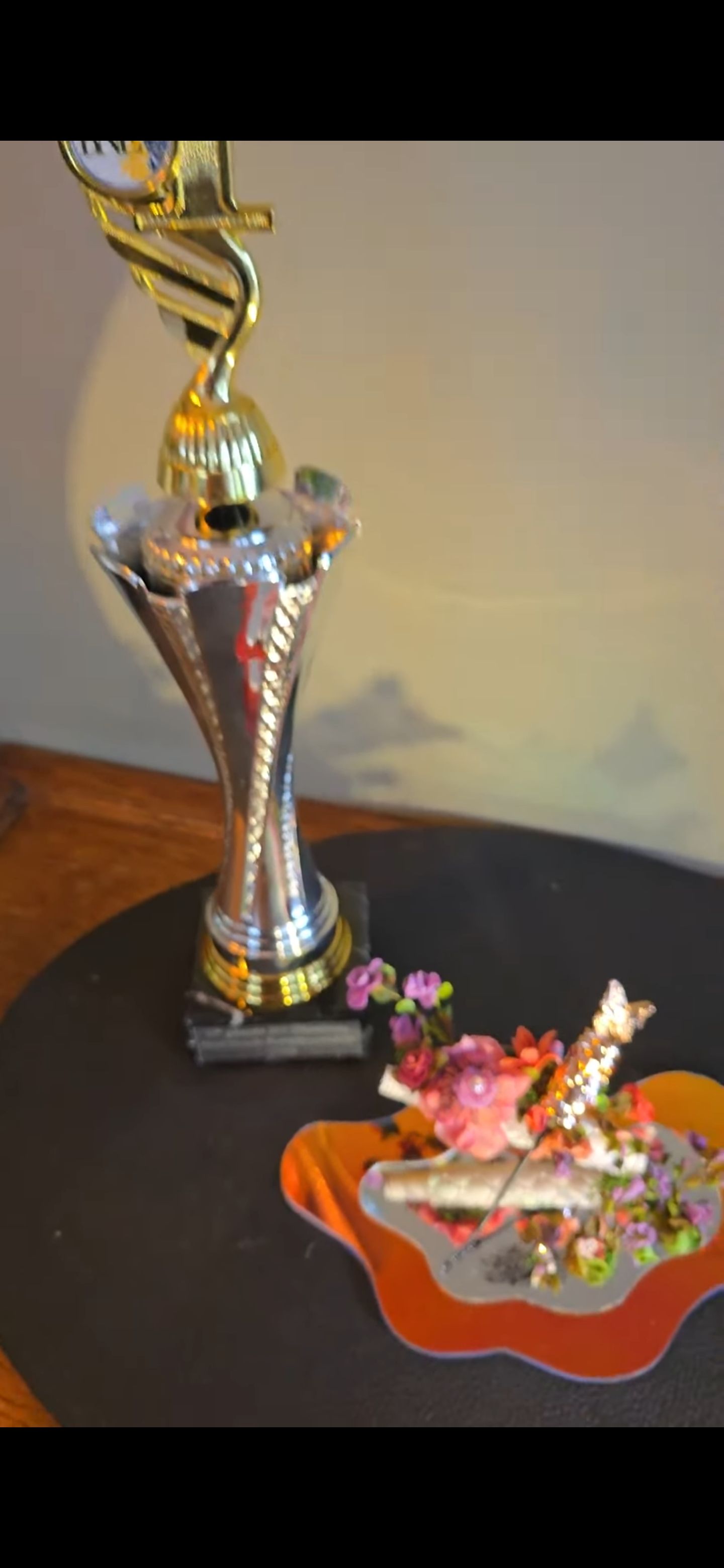 Naast een klein, spiegelend decoratief object met bloemen op een donkere ondergrond staat een zilveren en gouden trofee.