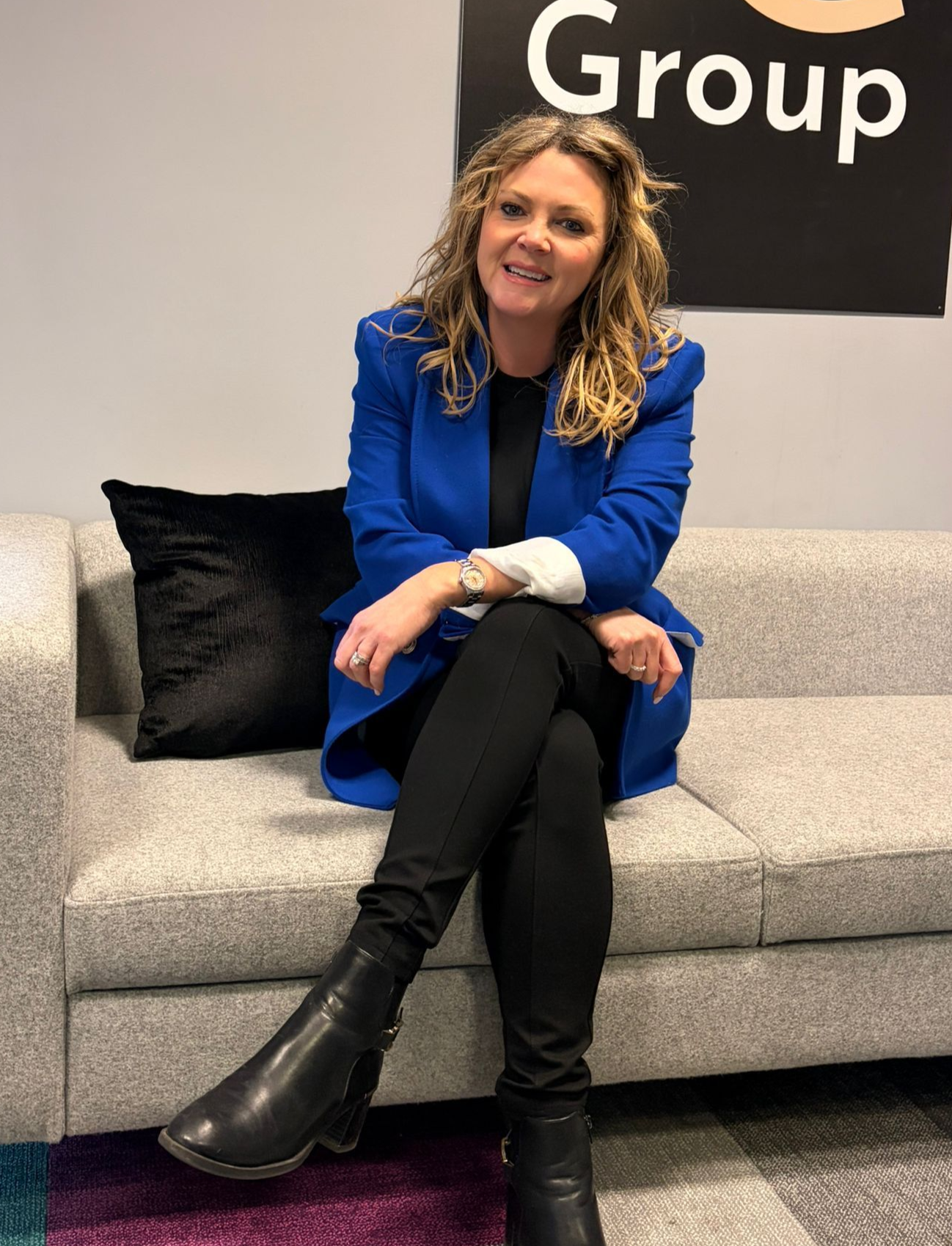Woman in a blue blazer seated on a gray couch in an office lounge, smiling at the camera.
