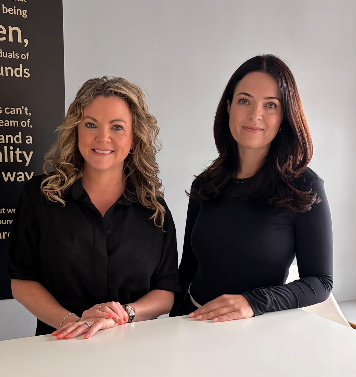 Two women in black tops smiling behind a white table in a bright office setting