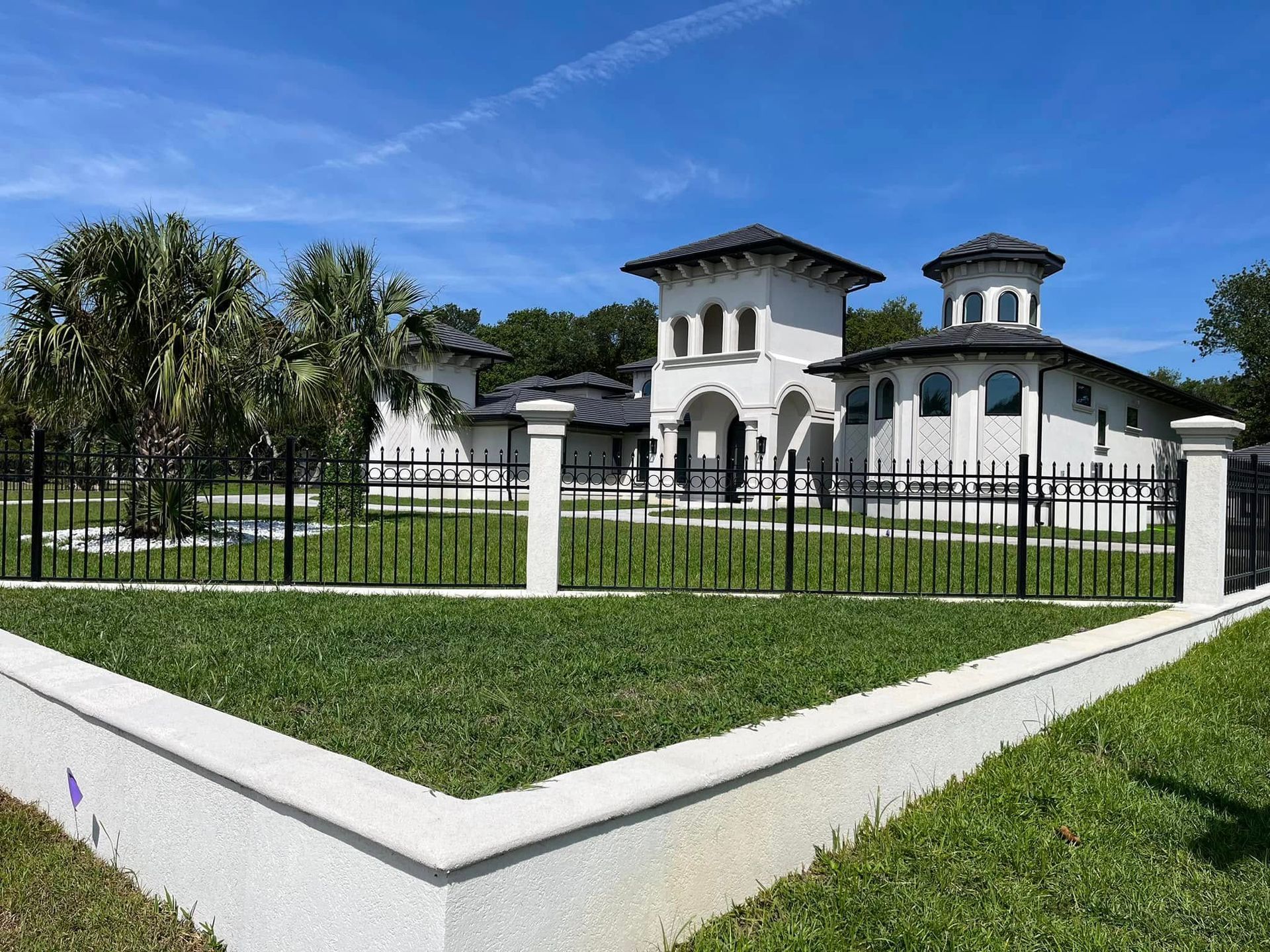 Large white mansion with a black fence and green lawn under a blue sky