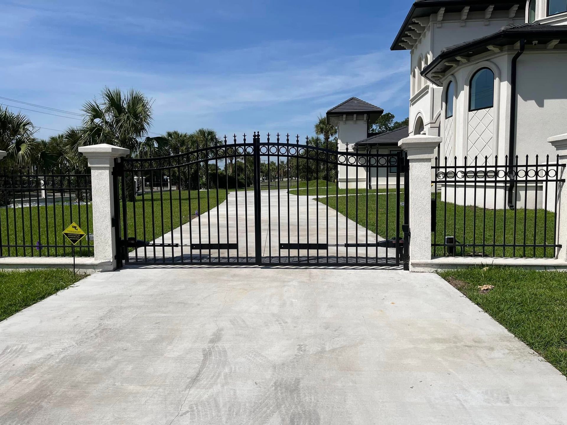 Black wrought-iron gate at the end of a concrete driveway, with a house and palm trees beyond.