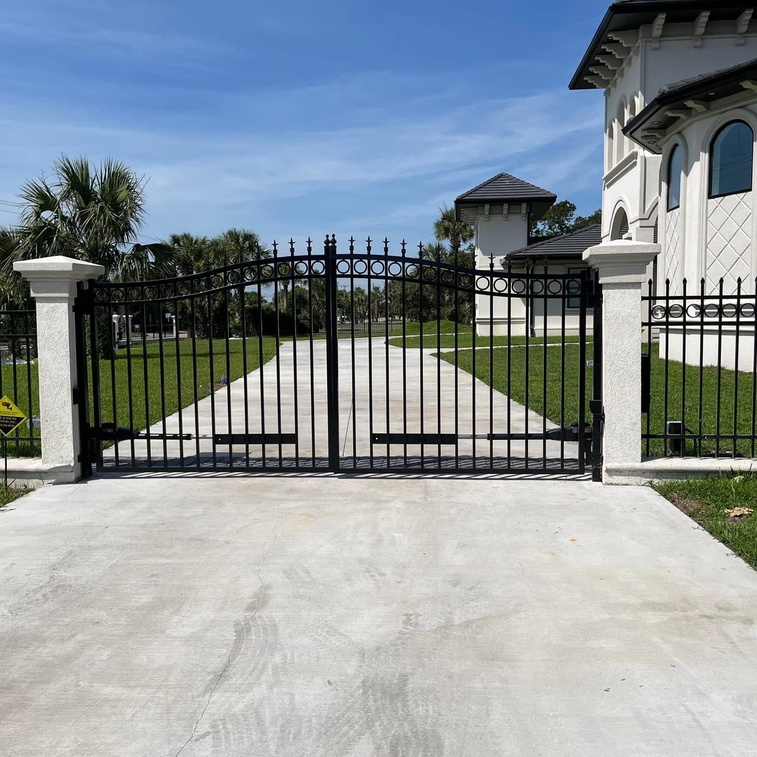 Black iron gate at the end of a concrete driveway beside a white house and green lawn under blue sky