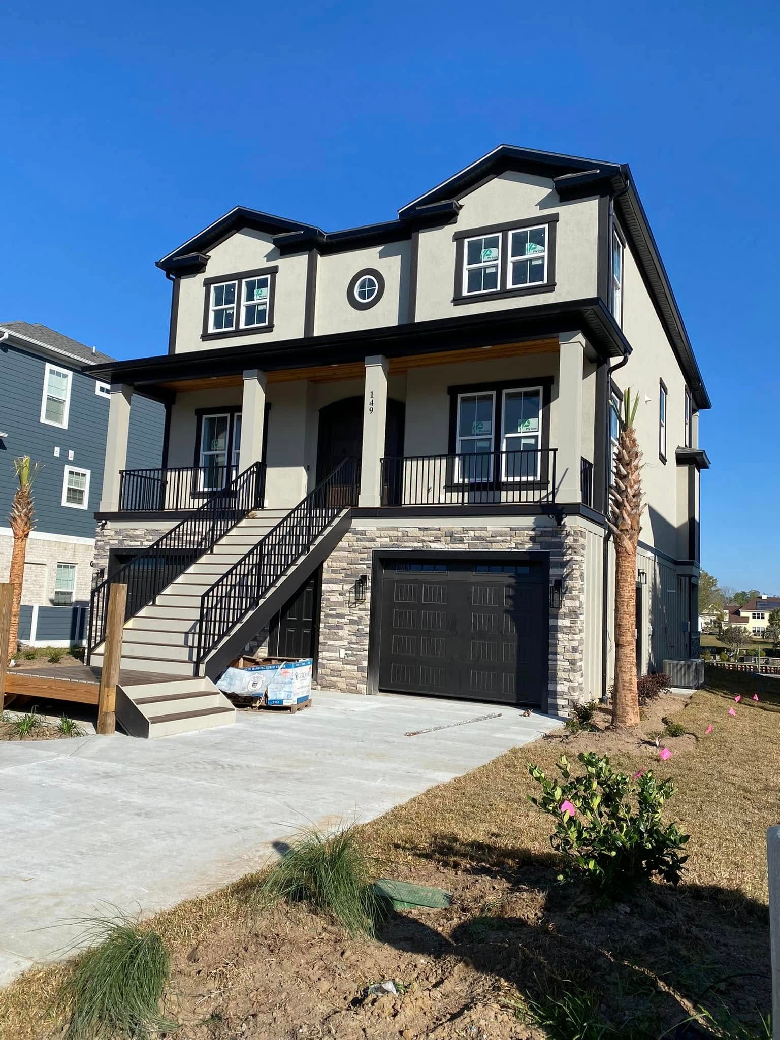 Two-story beige house with black trim, front porch, garage, and driveway under construction.