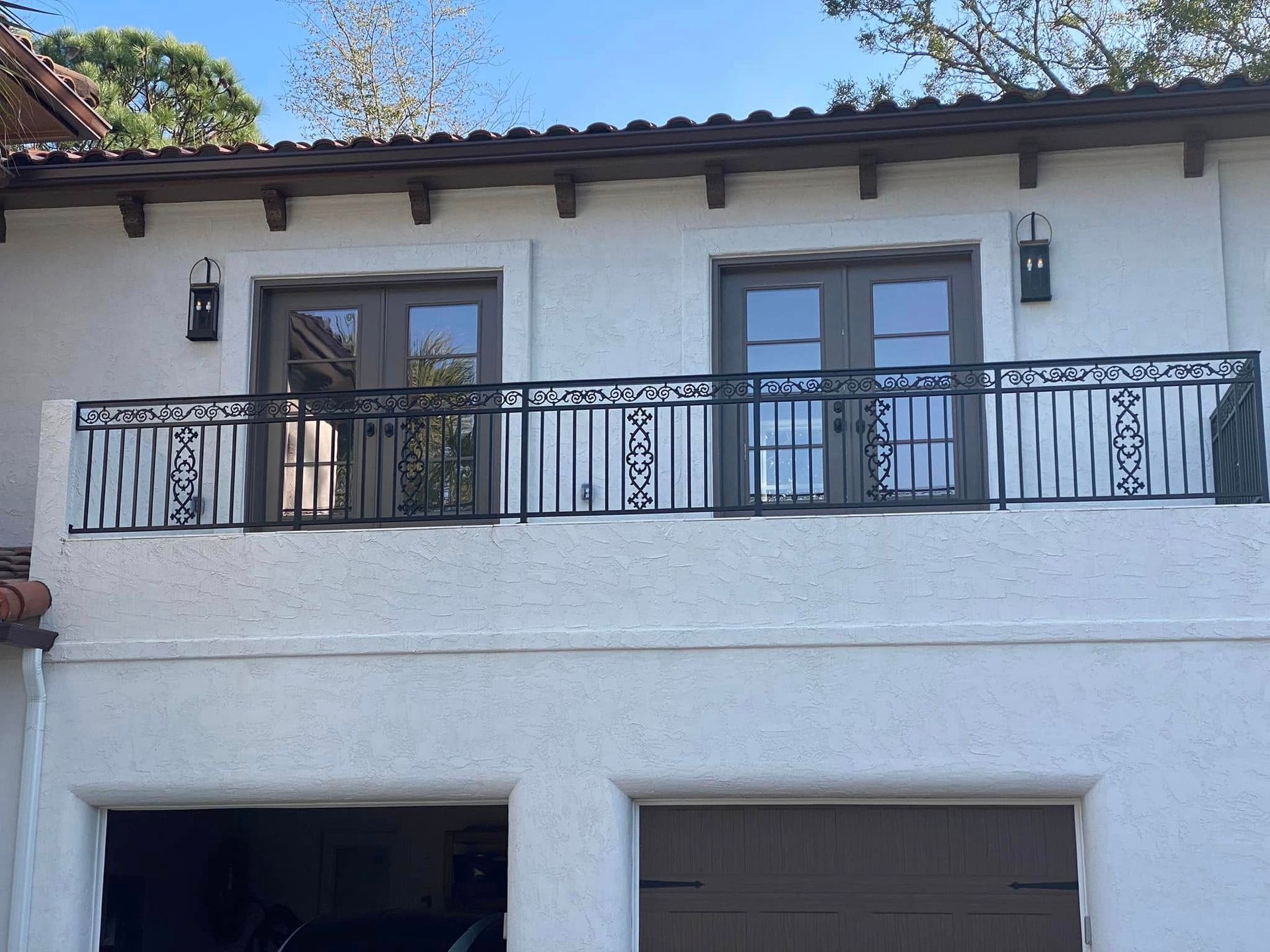 White stucco house facade with black wrought-iron balcony and two French doors above garage openings