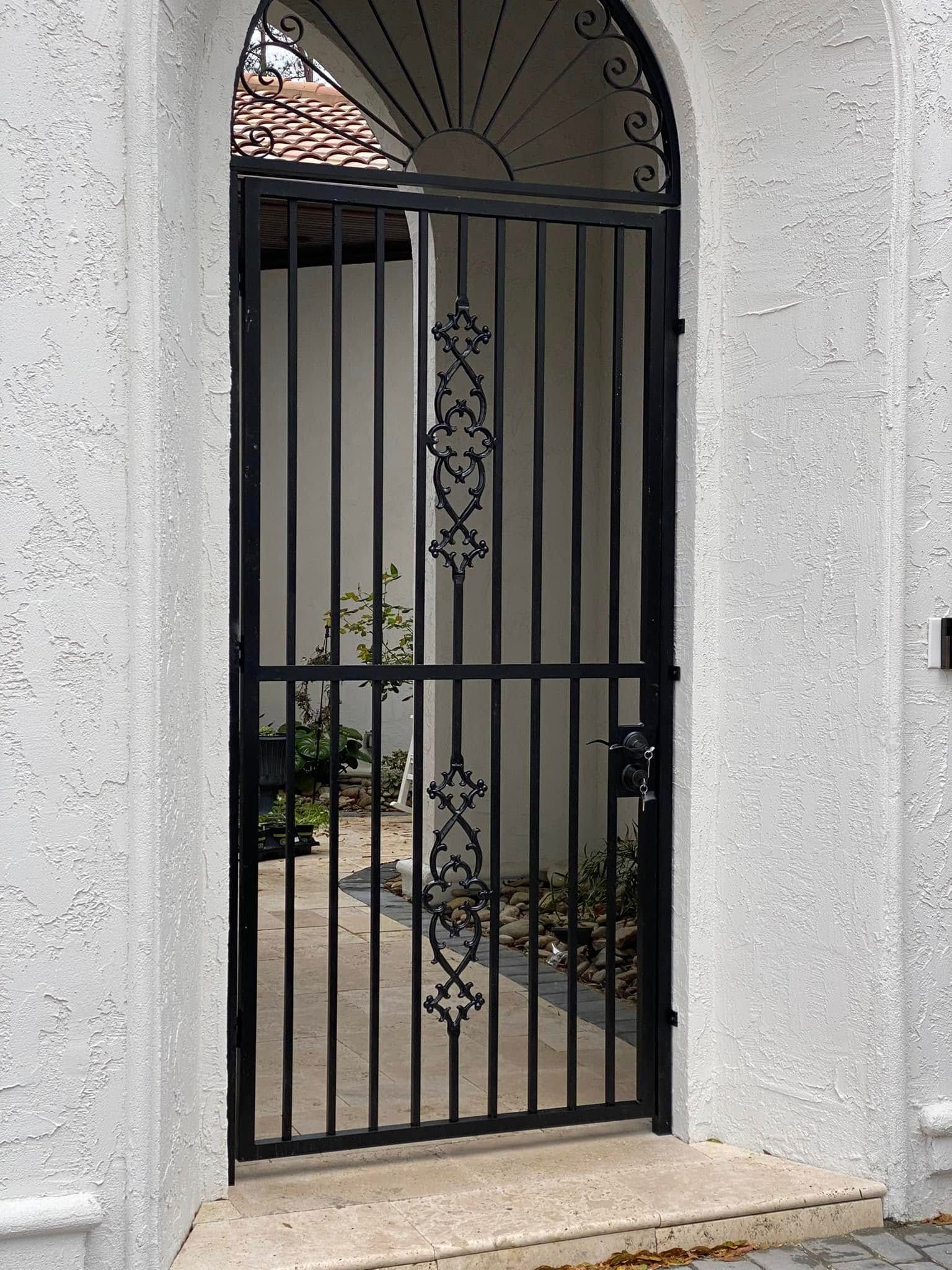 Black metal garden gate set in a white stucco wall, with decorative scrollwork and plants visible behind it