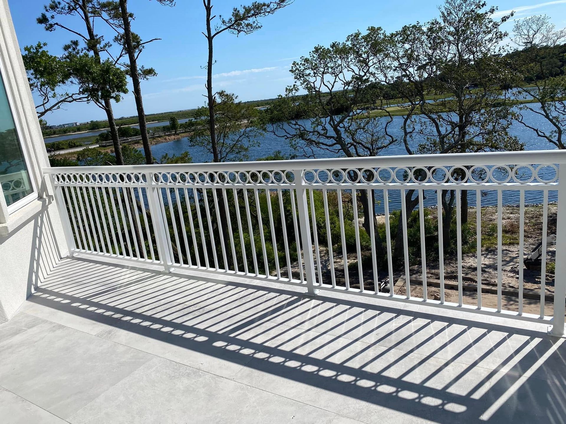 White balcony railing casting shadows beside a waterfront with trees and blue water under a clear sky