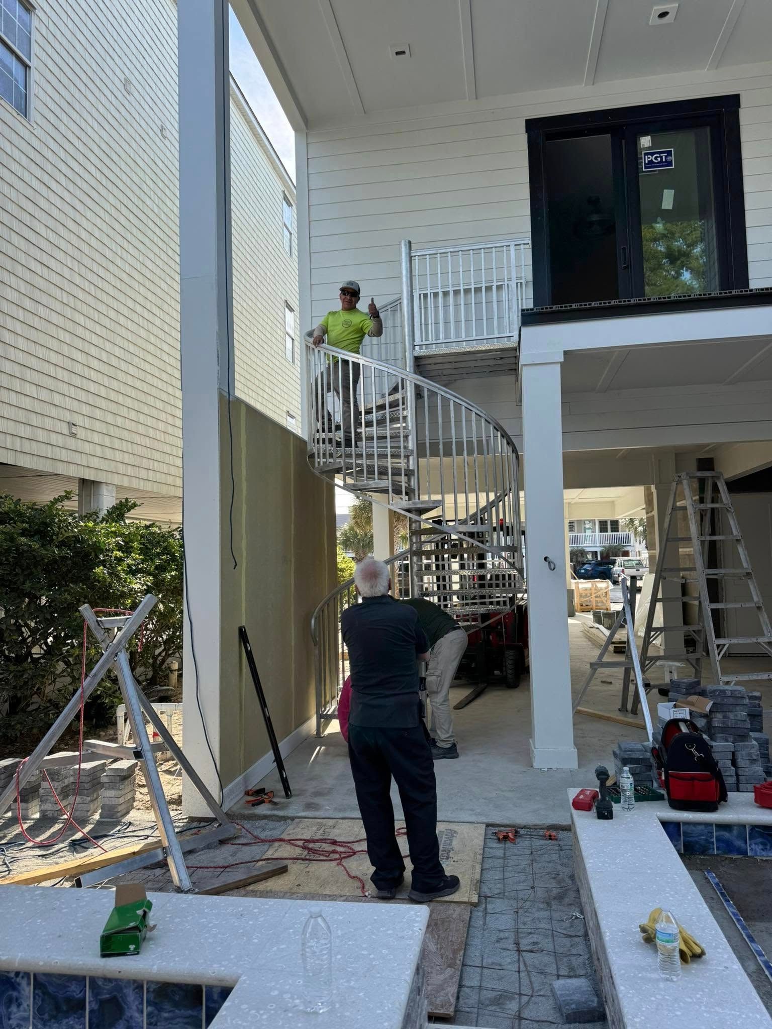 Workers on a house exterior, one on a spiral stair and another below, with tools and ladders around them