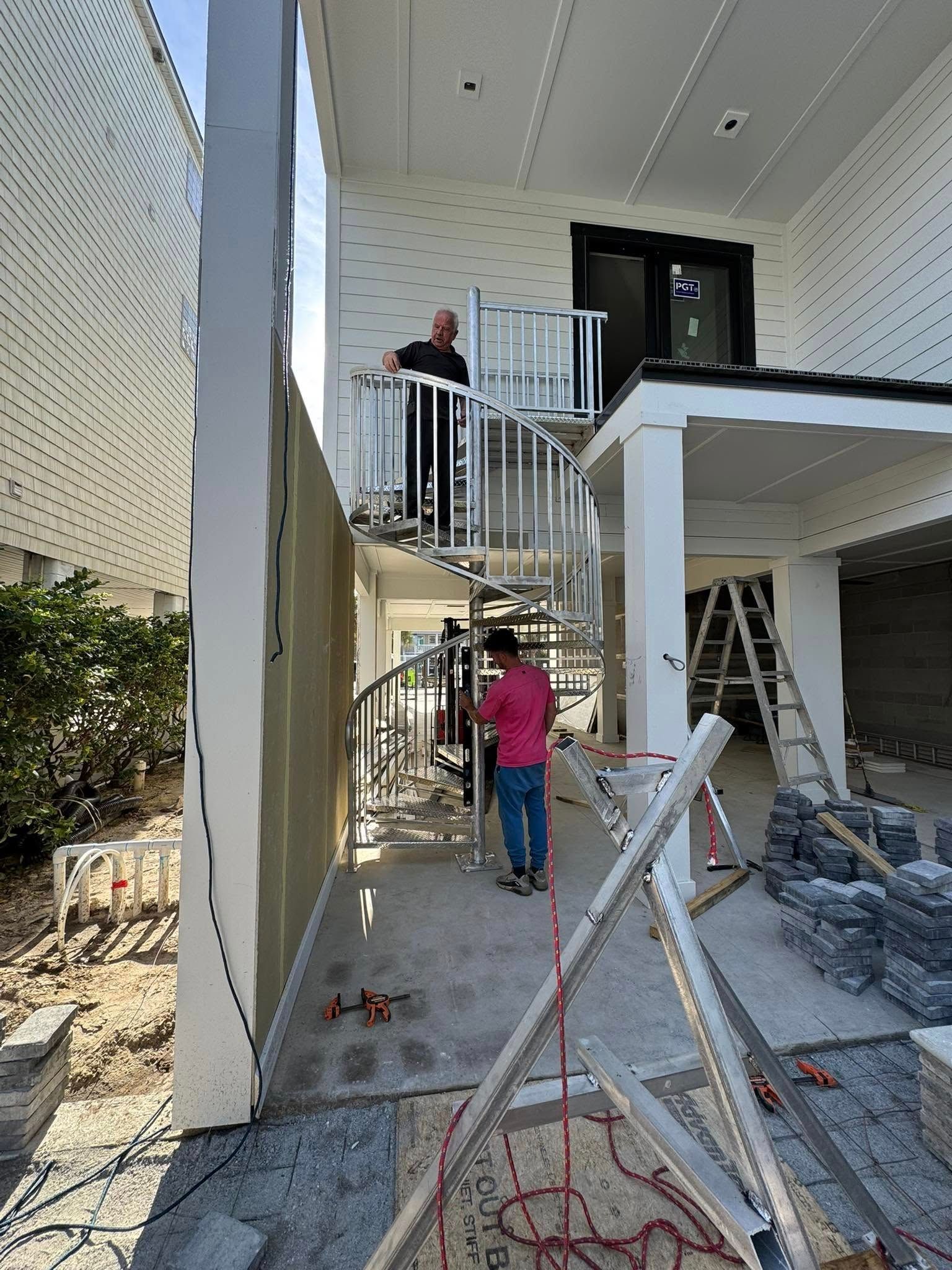 Two workers on a house exterior staircase under construction, with tools and materials scattered below.
