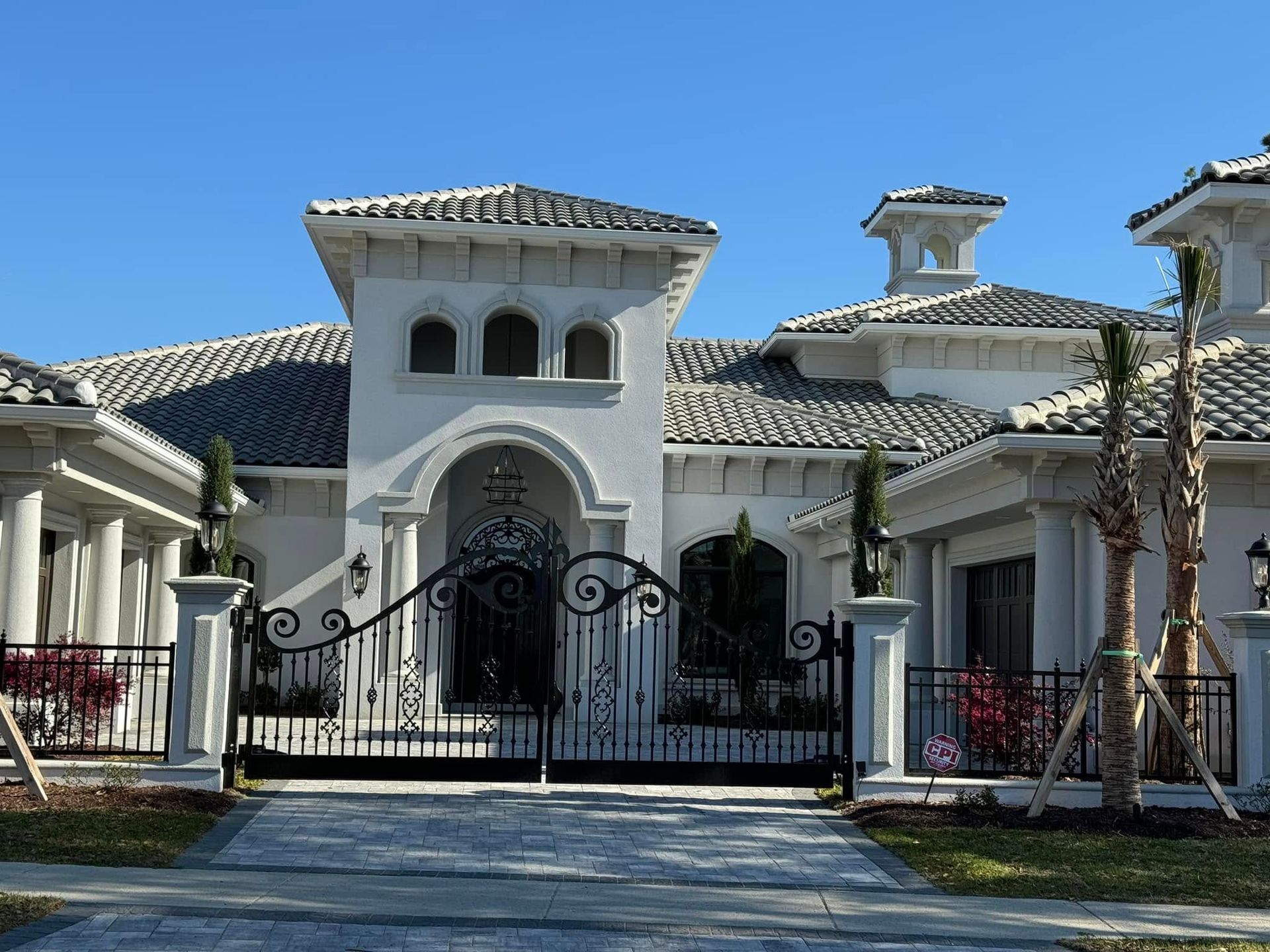 Large white Mediterranean-style house with a black wrought-iron gate and tiled roof under a clear blue sky