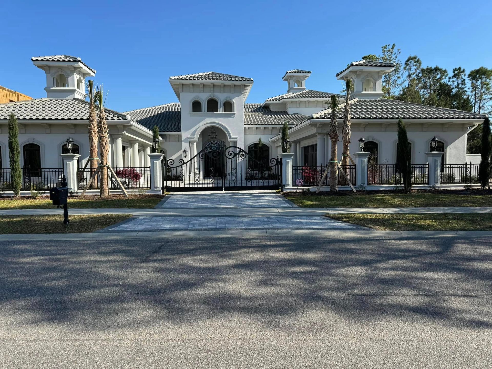 White Mediterranean-style mansion with black gates and a long driveway under a clear blue sky
