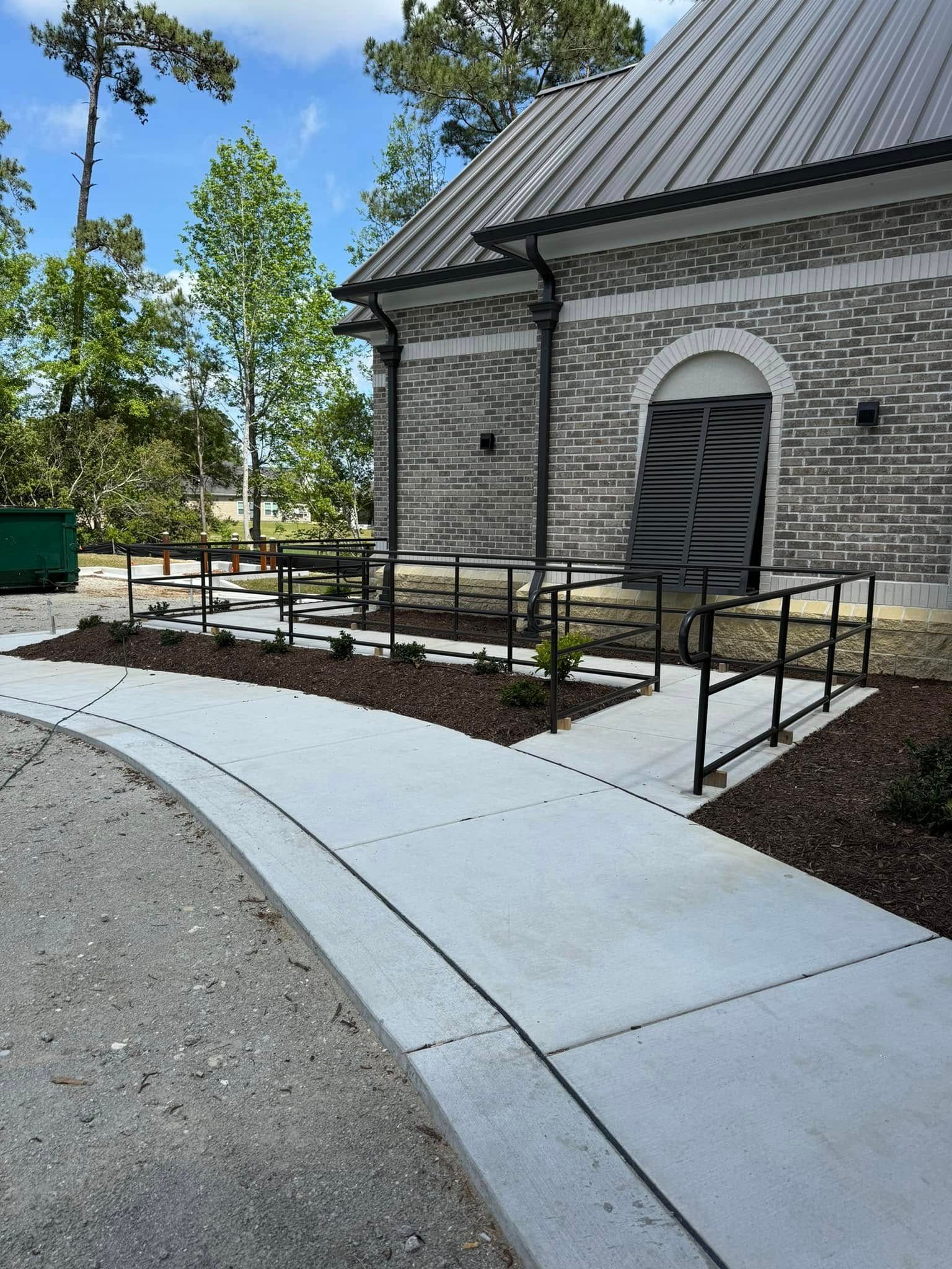 Curved concrete sidewalk leading to a stone building entrance with black railings and landscaping
