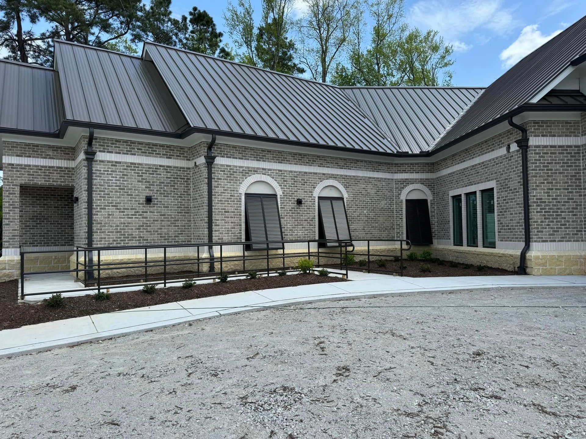 Stone building with metal roof, arched windows, and ramp along a gravel driveway.