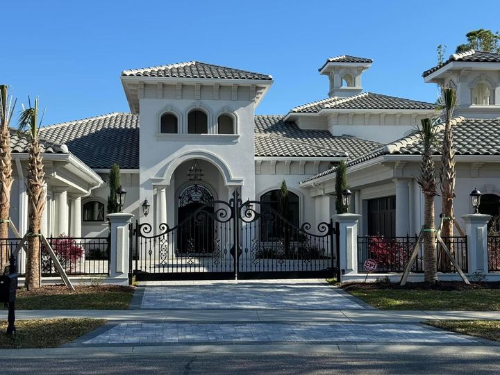 Gated Mediterranean-style mansion with white stucco walls, arched entry, and tiled roofs under a blue sky
