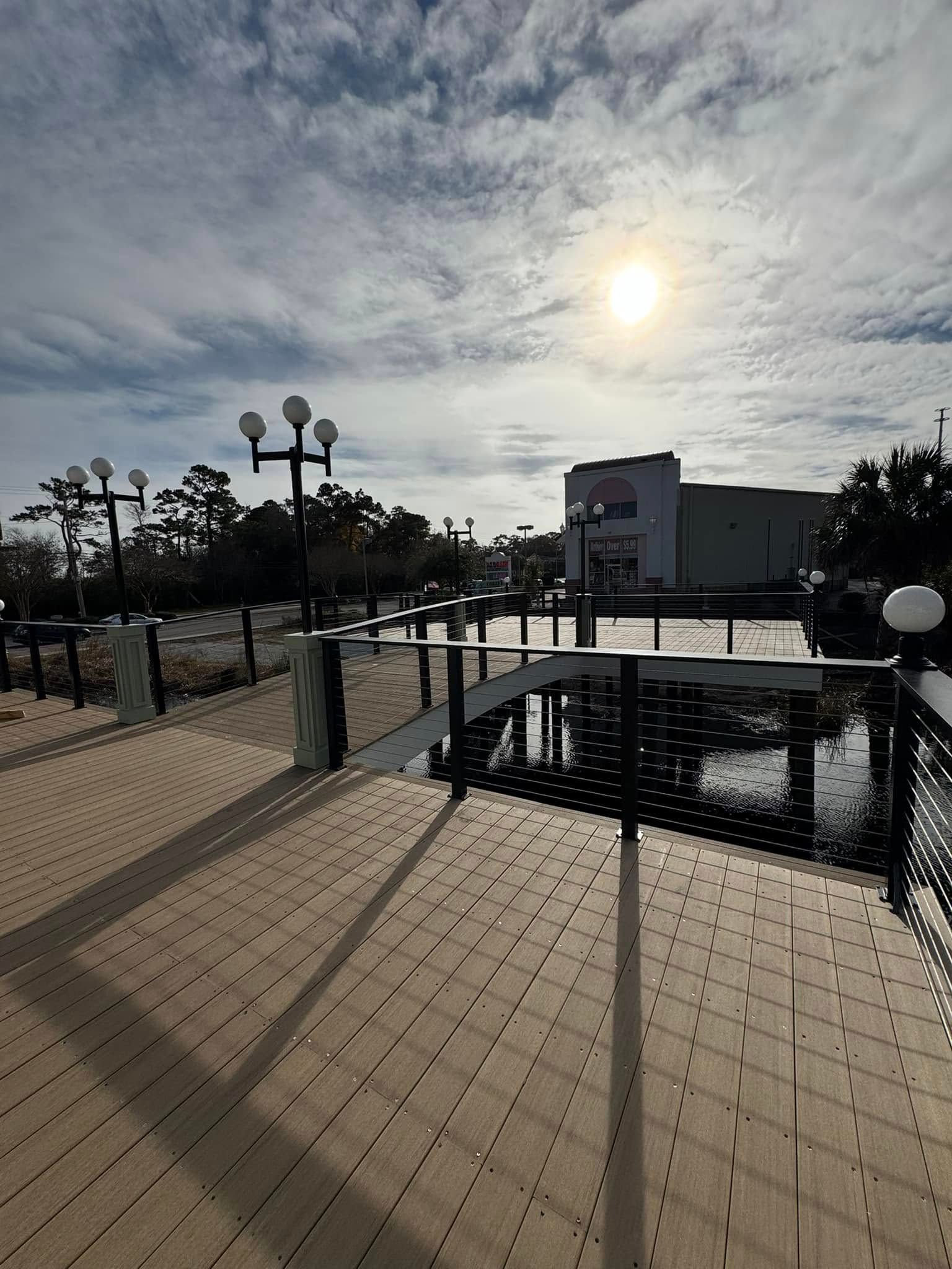 Sunlit outdoor deck with metal railings, tiled flooring, lamp posts, and cloudy sky over water
