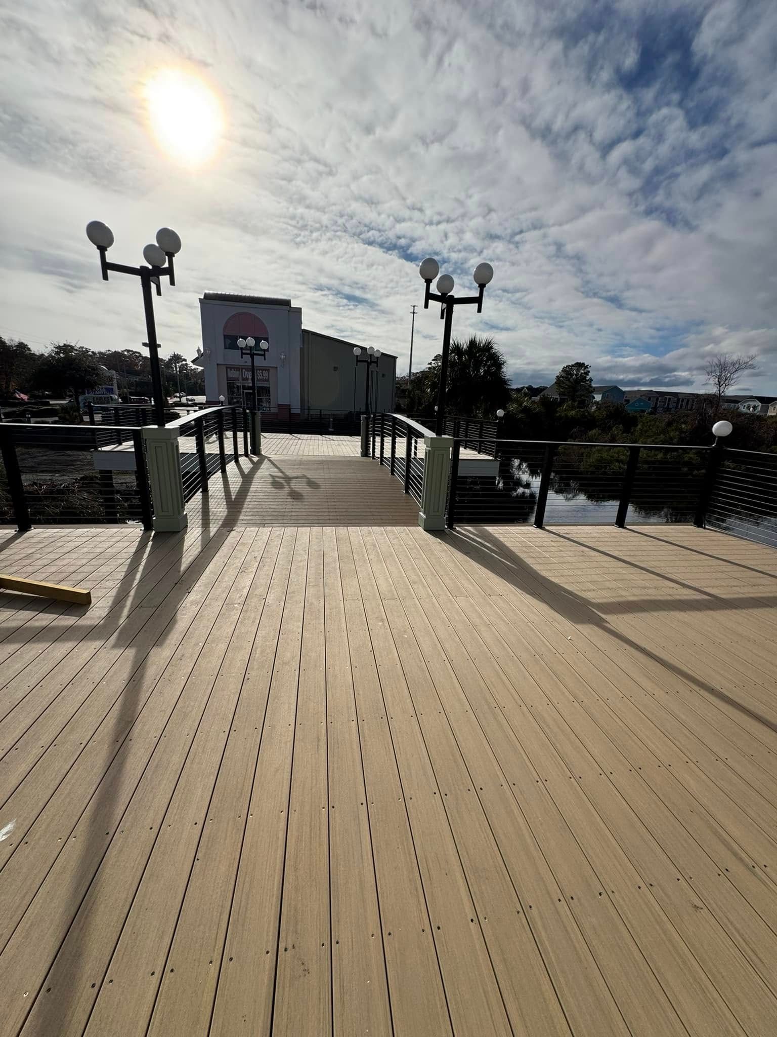 Sunlit wooden boardwalk leading to a building, with lamp posts and water on both sides under a cloudy sky
