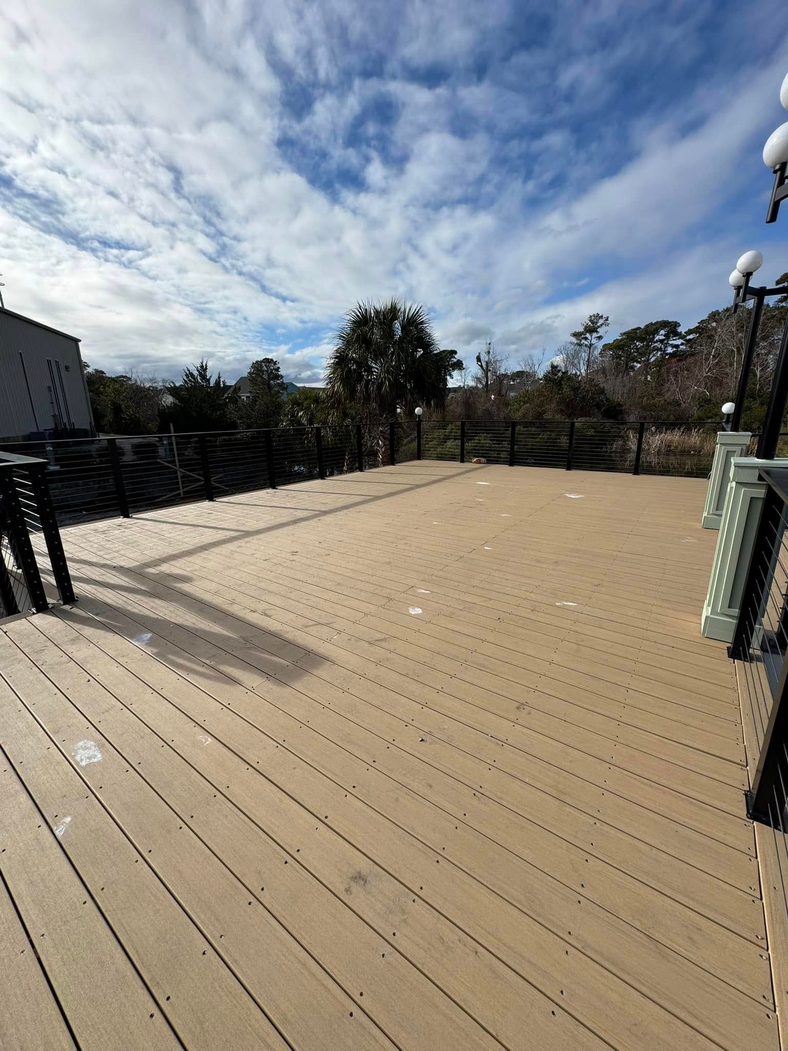 Wide tan wooden deck under a cloudy blue sky, with trees and black railings around it.