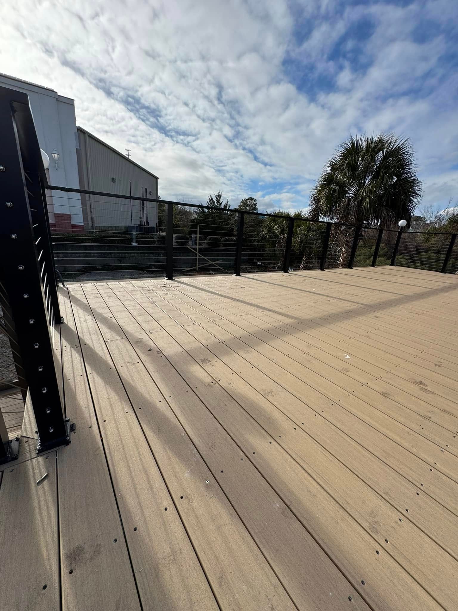 Wooden deck with metal railing beside a building, overlooking trees under a partly cloudy sky