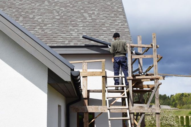 Worker on a ladder repairing a roof beside a house under a cloudy sky