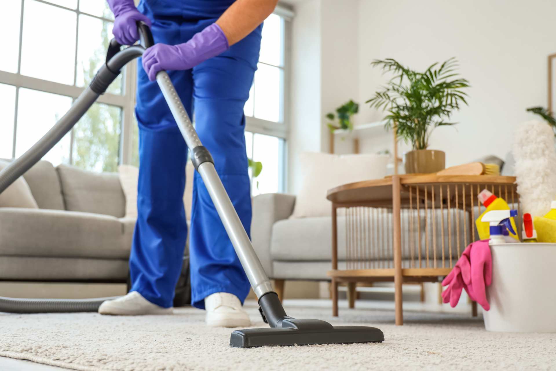Person vacuuming a carpet in a bright living room with a gray sofa and wooden table