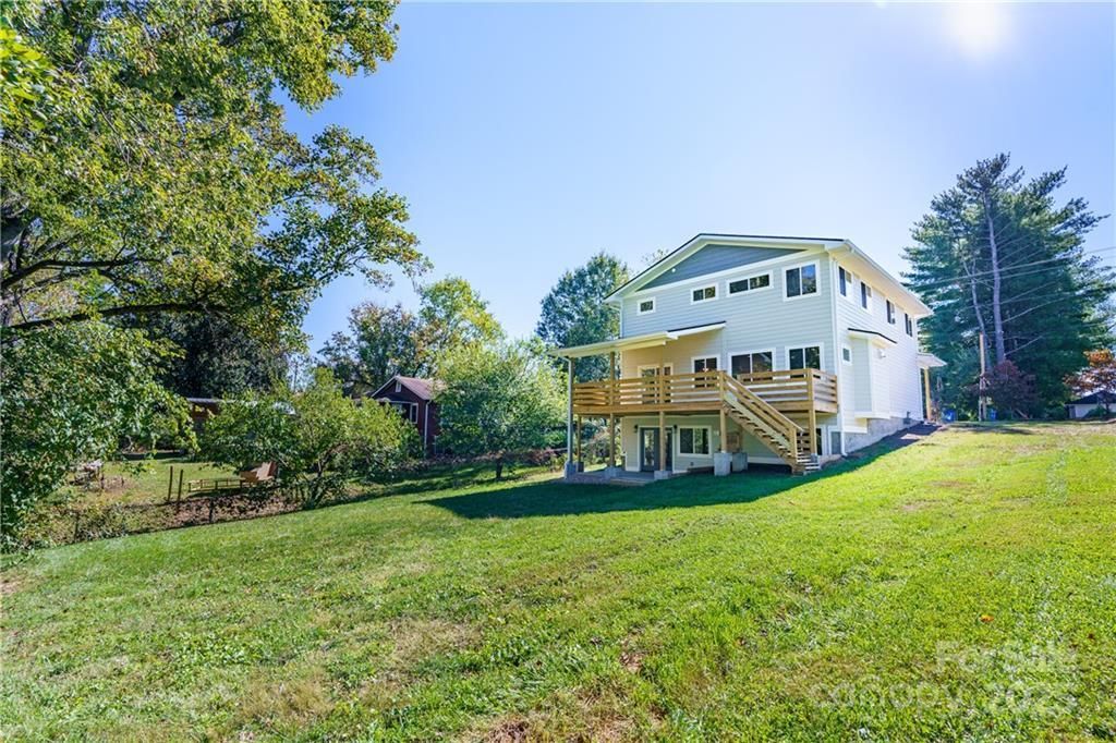 Large white house with a wooden deck, situated on a grassy hill with trees and a clear blue sky.