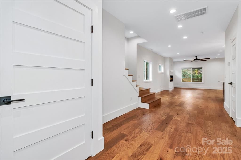 Bright empty living room with hardwood floors, white walls, stairs, and large windows.