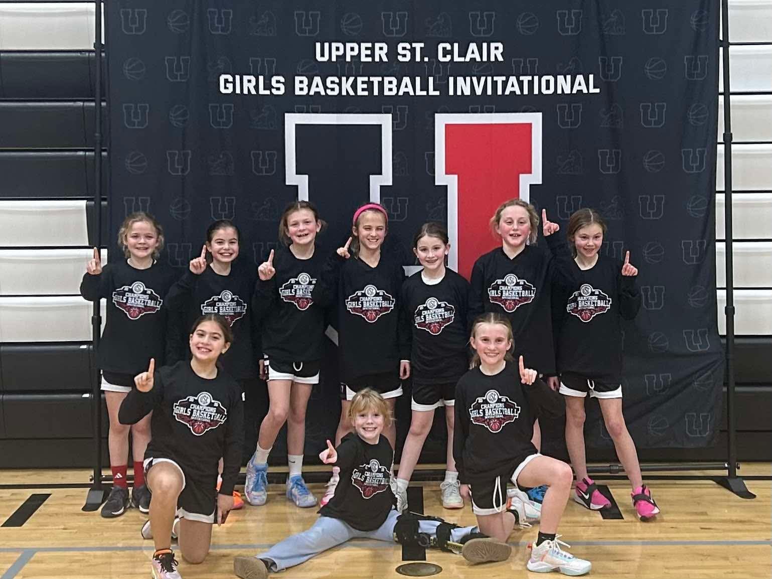 Girls basketball team posing in black uniforms at the Upper St. Clair Invitational backdrop
